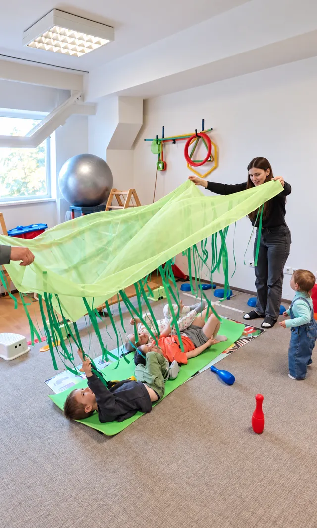 Специалист и взрослый держат зелёную ткань над детьми во время групповой игры